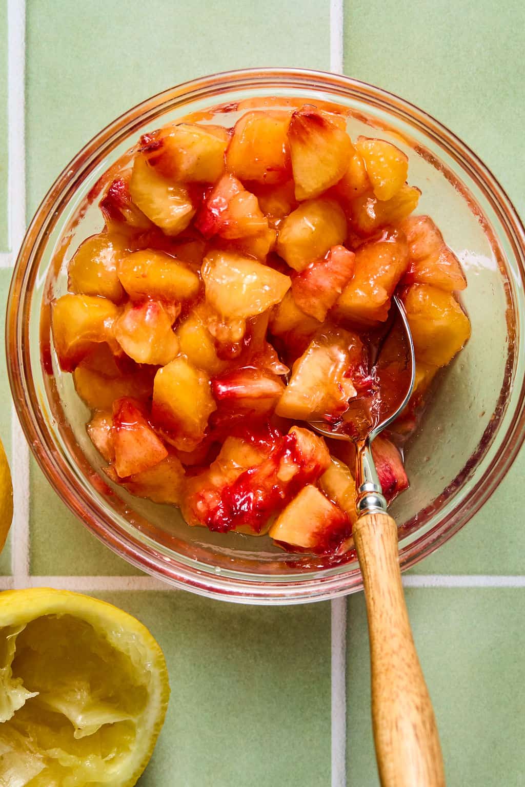 A glass bowl filled with chopped peaches in syrup sits on a green tiled surface. A metal spoon with a wooden handle rests inside the bowl. Part of a squeezed lemon is visible in the lower left corner.