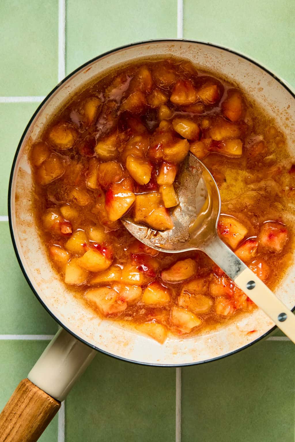 A saucepan with chunky peach and red fruit jam sits on a green tiled surface. A spoon rests in the pot, partially submerged in the golden, bubbling mixture.