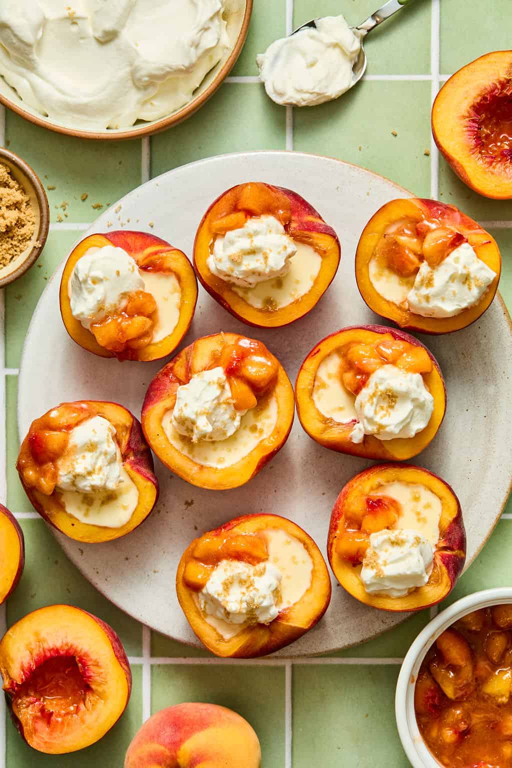 Halved peaches on a plate topped with ice cream, peach compote, and a drizzle of sauce. Surrounding the plate are bowls of whipped cream, brown sugar, and whole peaches on a tiled green surface.
