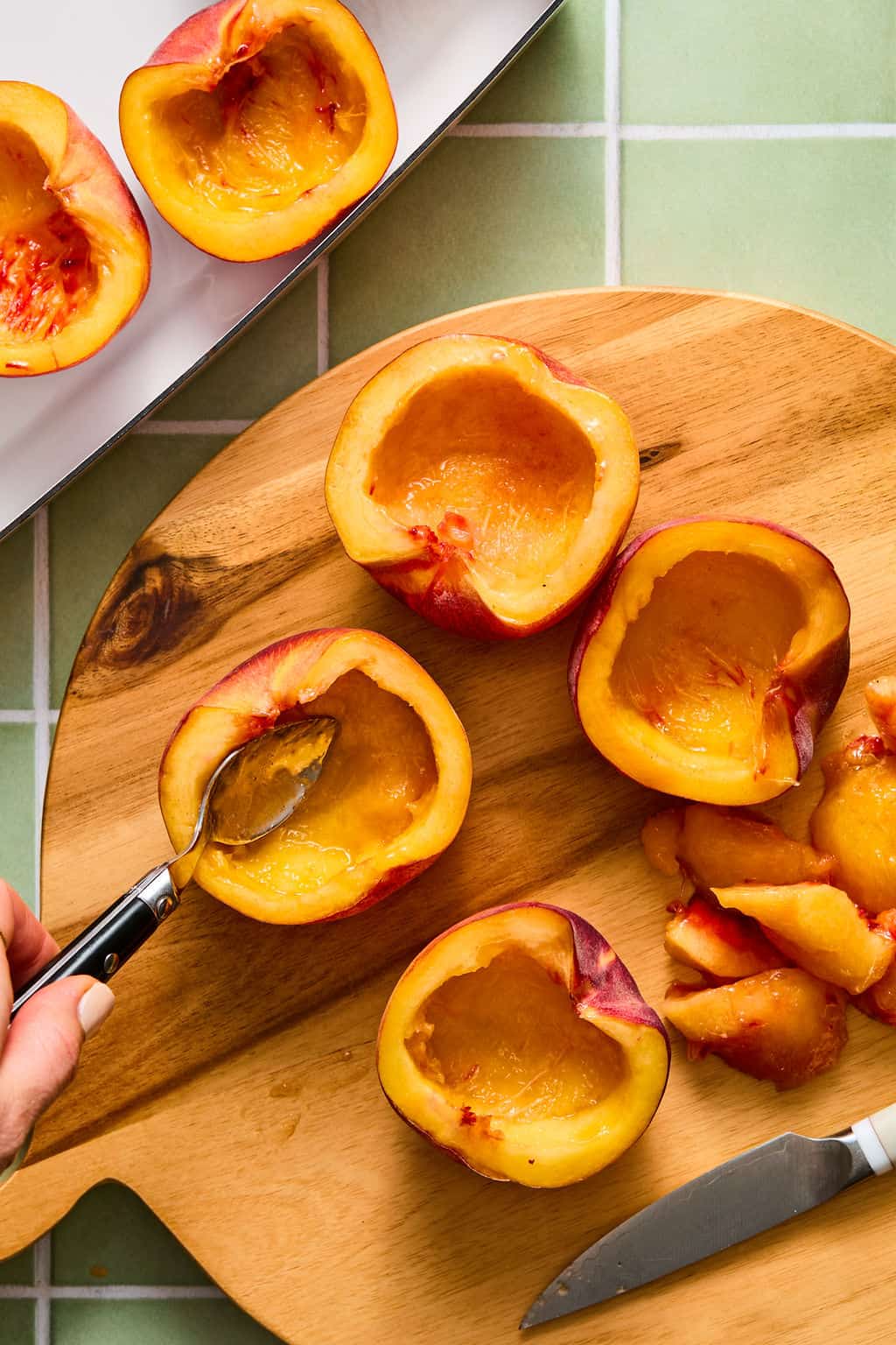 Halved peaches with pits removed are on a wooden cutting board. One peach is being hollowed with a spoon. A paring knife, peach slices, and more peach halves are nearby on a green tiled surface.