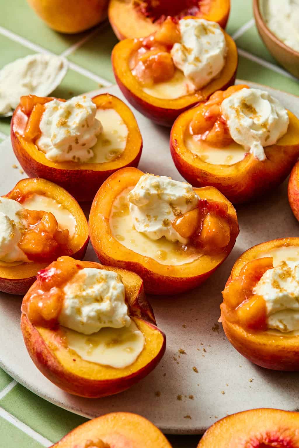 Halved peaches topped with whipped cream, a creamy sauce, and fruit compote are arranged on a plate. The dessert sits on a green and white checkered tablecloth.
