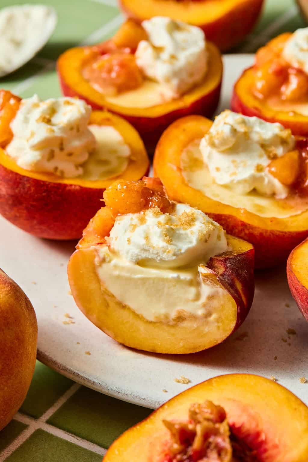 Halved peaches topped with whipped cream and a sprinkle of brown sugar are arranged on a plate. The peaches sit on a green and white tiled surface, creating a fresh, summery presentation.