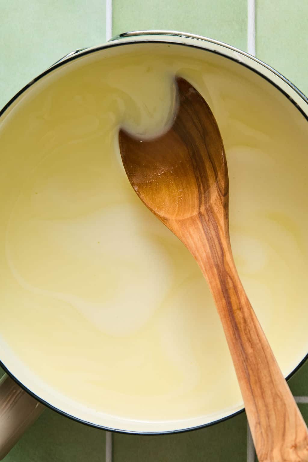 A wooden spoon stirs a creamy white sauce in a pot, set on a green tiled surface.