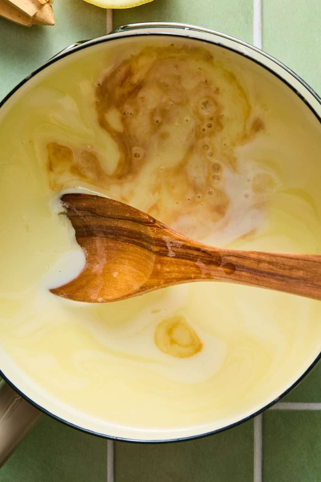 A wooden spoon stirs a creamy yellow mixture with brown vanilla extract swirls in a white pot on a green tiled surface.