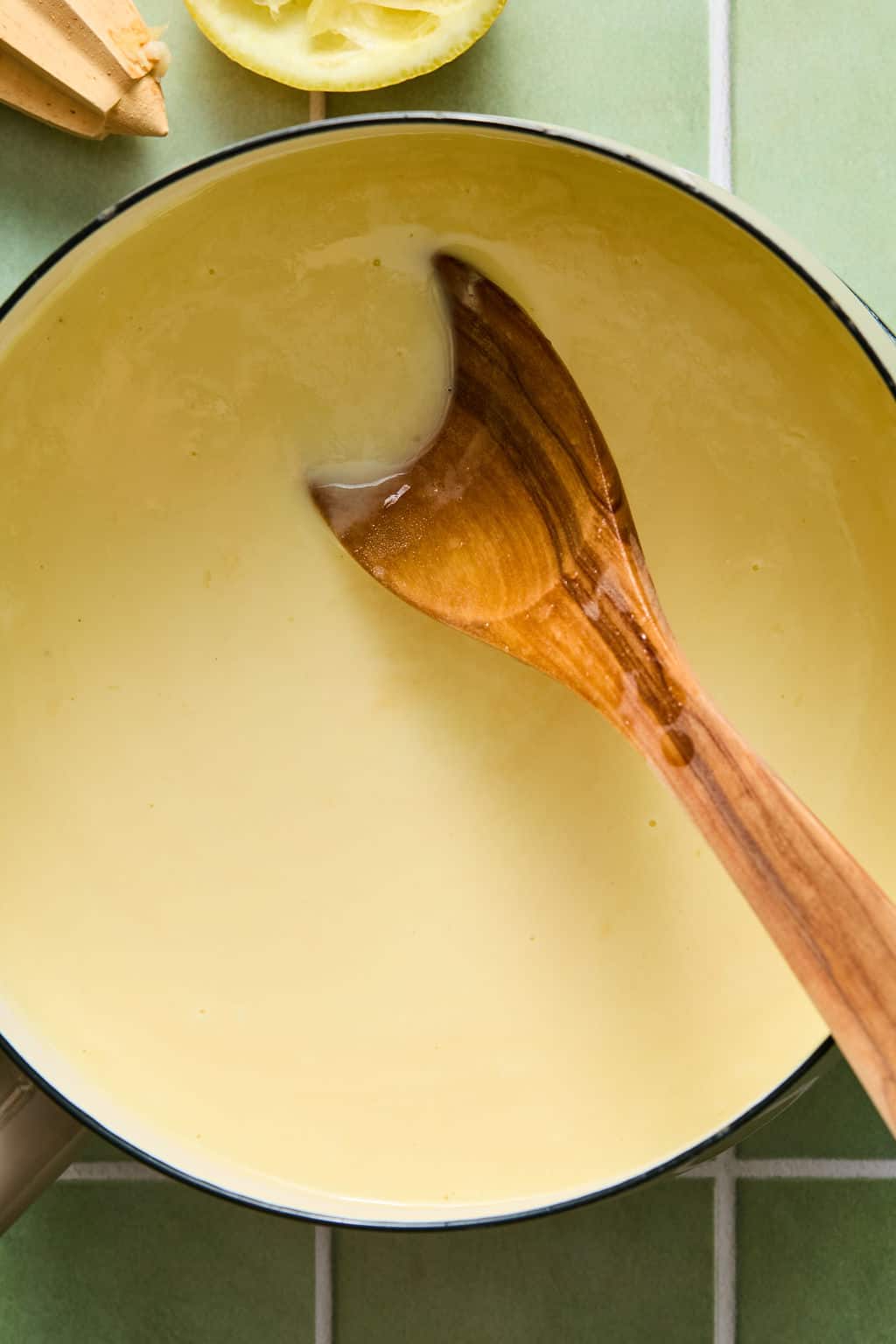 A wooden spoon stirs a creamy yellow sauce in a white saucepan on a green tiled surface, with a lemon half and juicer partially visible in the corner.