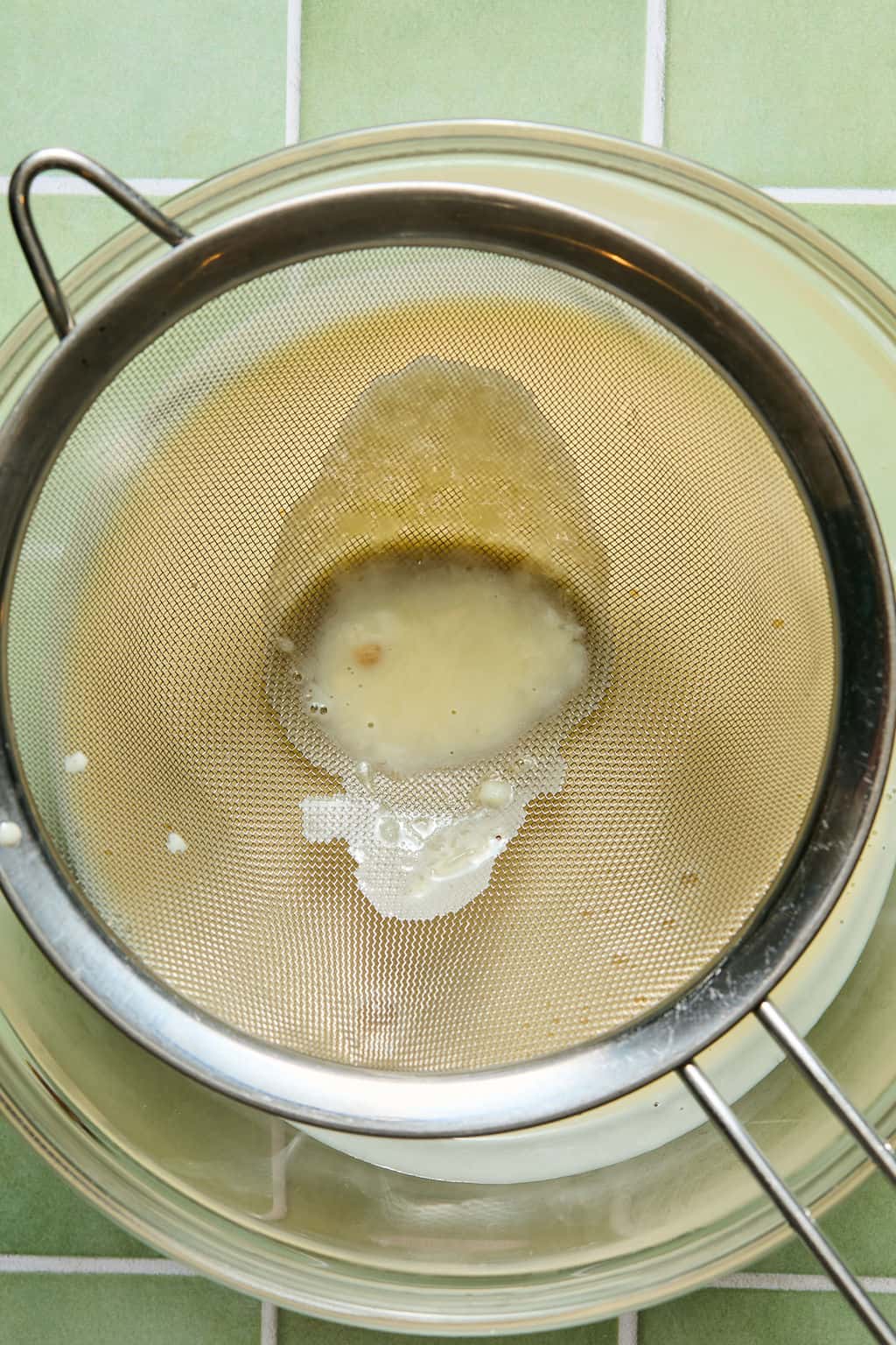 A metal strainer over a glass bowl holds pulp while liquid drains through, set on a green tiled surface.