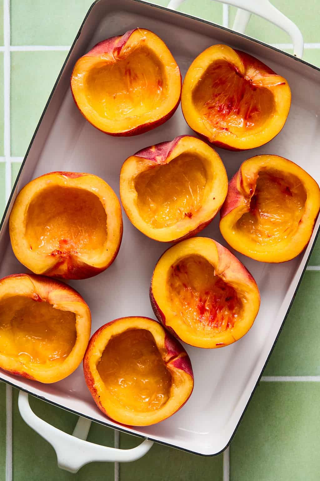 Eight halved and pitted peaches arranged cut side up in a white rectangular baking dish on a light green tiled surface.