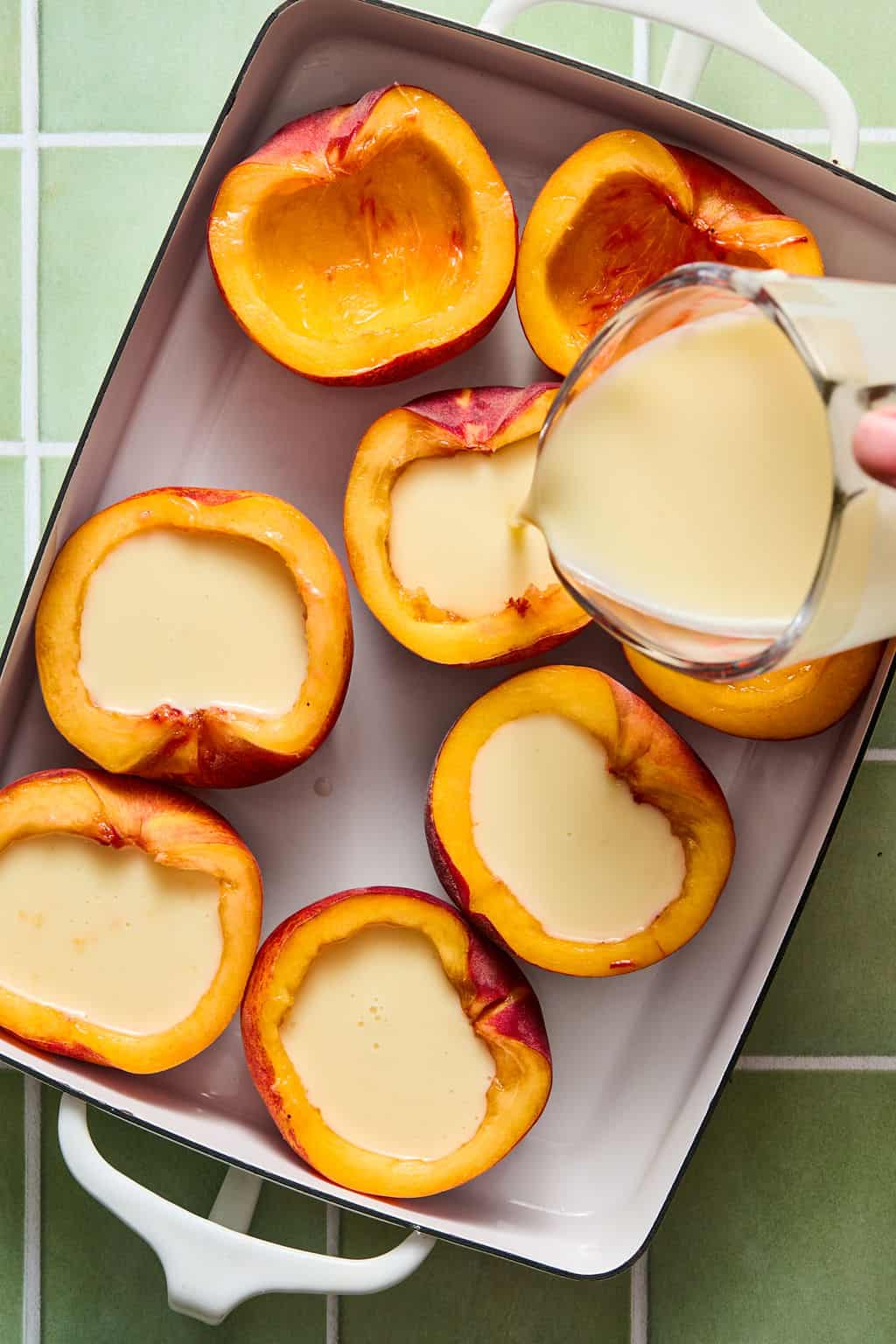 Halved peaches in a white baking dish with custard being poured into the centers. The background is a green and white tiled surface.