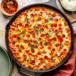 A cast iron skillet filled with cheesy pepperoni pizza orzo, garnished with herbs, with a small bowl of chili oil and grated cheese nearby.