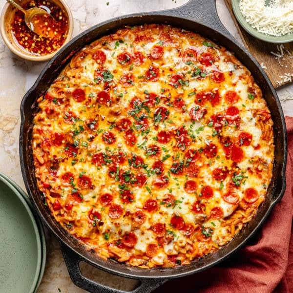 A cast iron skillet filled with cheesy pepperoni pizza orzo, garnished with herbs, with a small bowl of chili oil and grated cheese nearby.