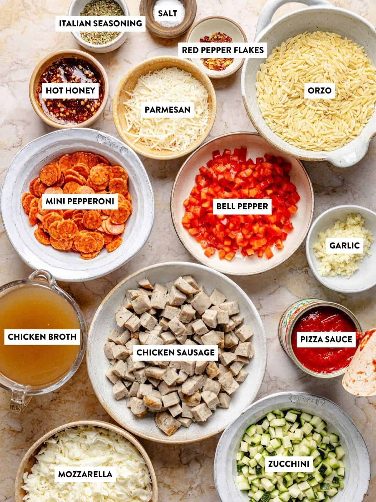 Bowls of labeled ingredients for a pizza orzo recipe, including Italian seasoning, hot honey, parmesan, mini pepperoni, bell pepper, garlic, chicken broth, chicken sausage, pizza sauce, mozzarella, and zucchini.