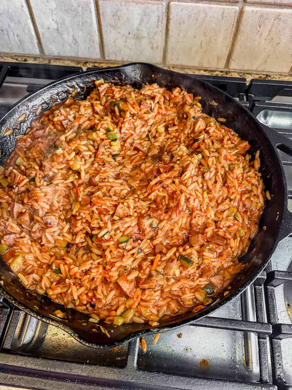 Cast iron skillet filled with cooked orzo pasta in a tomato-based sauce with herbs, sitting on a stove with a tiled backsplash in the background.
