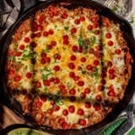 A cast iron skillet filled with a baked pizza orzo topped with melted cheese, mini pepperoni slices, and sprinkled herbs. Sunlight highlights parts of the golden, cheesy surface.