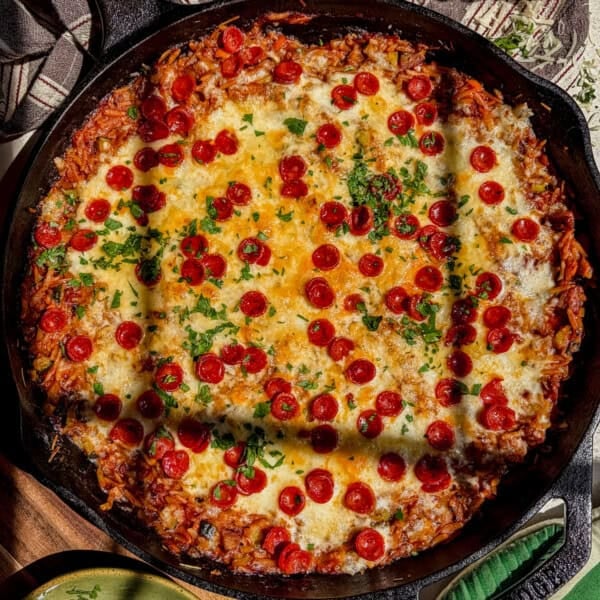 A cast iron skillet filled with a baked pizza orzo topped with melted cheese, mini pepperoni slices, and sprinkled herbs. Sunlight highlights parts of the golden, cheesy surface.