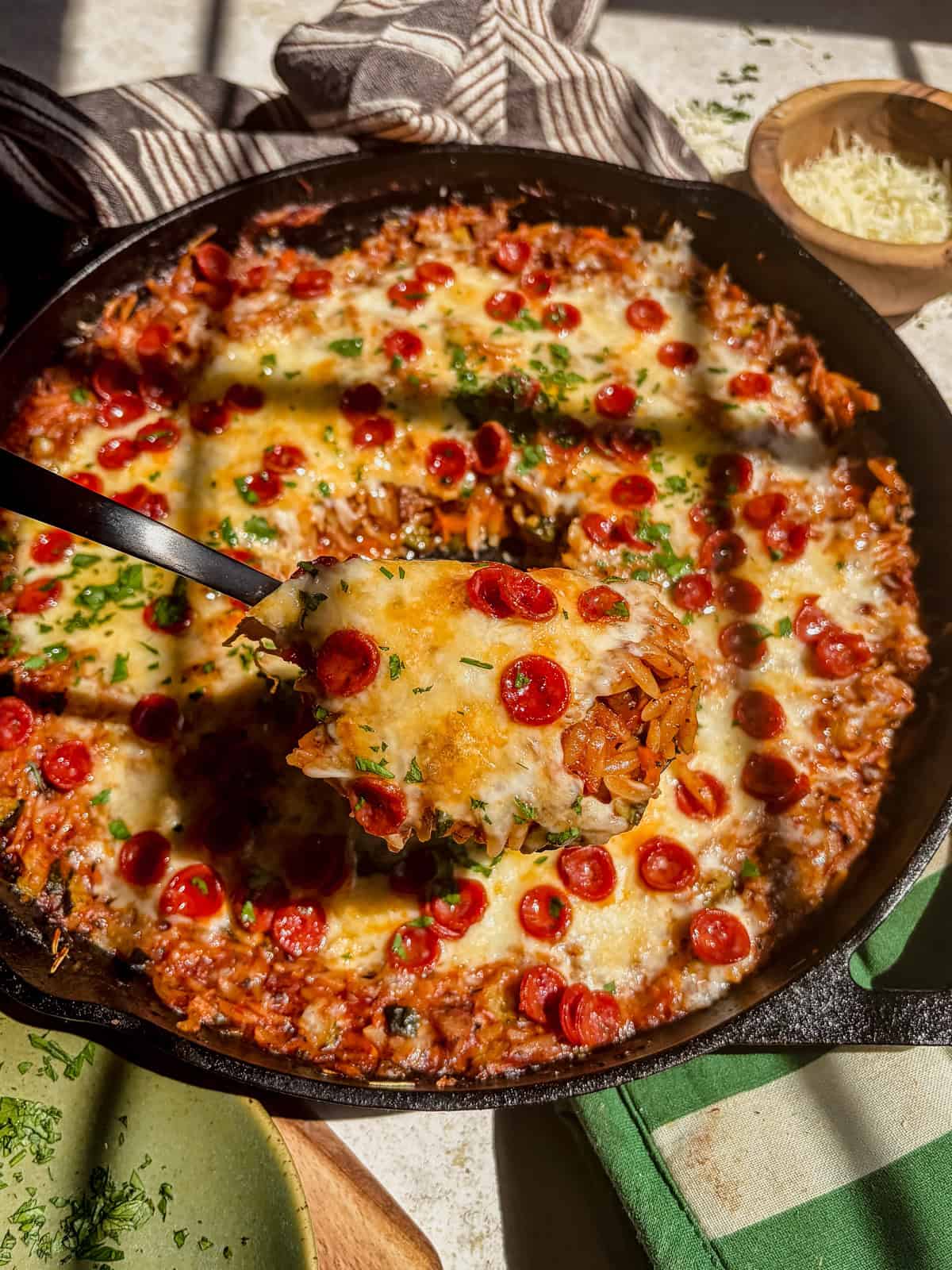 A skillet filled with cheesy baked pizza orzo topped with mini pepperoni slices and herbs; a serving is being lifted with a spatula, showing the melty cheese and pasta layers.