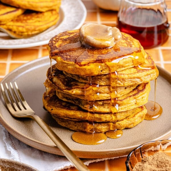A stack of fluffy pumpkin spiced pancakes topped with cinnamon butter and syrup sits on a plate with a gold fork, surrounded by more pancakes, syrup, and cinnamon on a tiled surface.