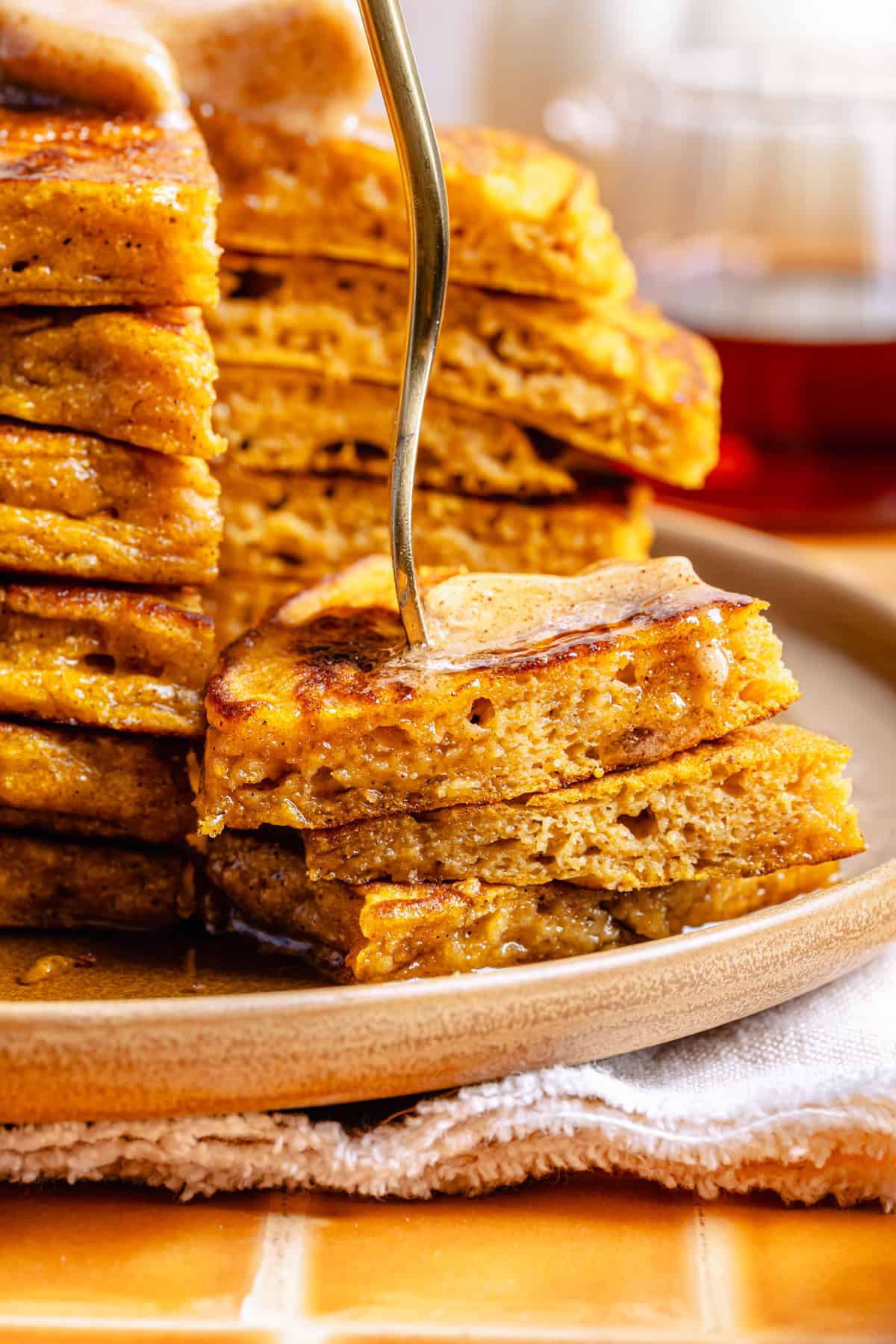A stack of thick, fluffy pumpkin spice pancakes with a pat of melting butter, syrup drizzled on top, and a fork cutting through the stack on a beige plate, sitting on a cloth napkin.