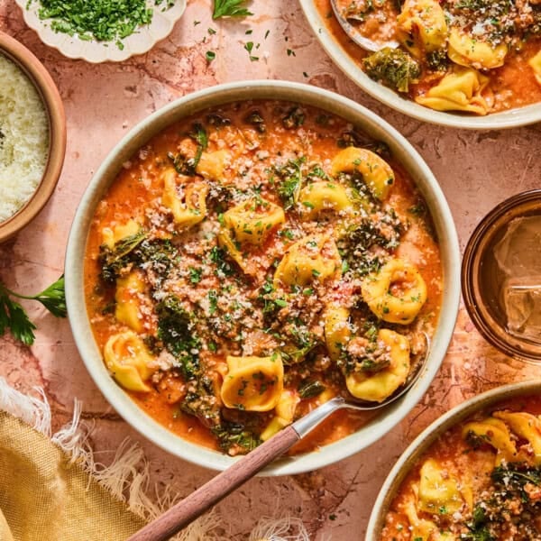 A top-down view of bowls of creamy sausage and tortellini soup with kale, topped with parmesan cheese and fresh herbs, arranged on a textured pink surface with extra cheese and a glass of water nearby.