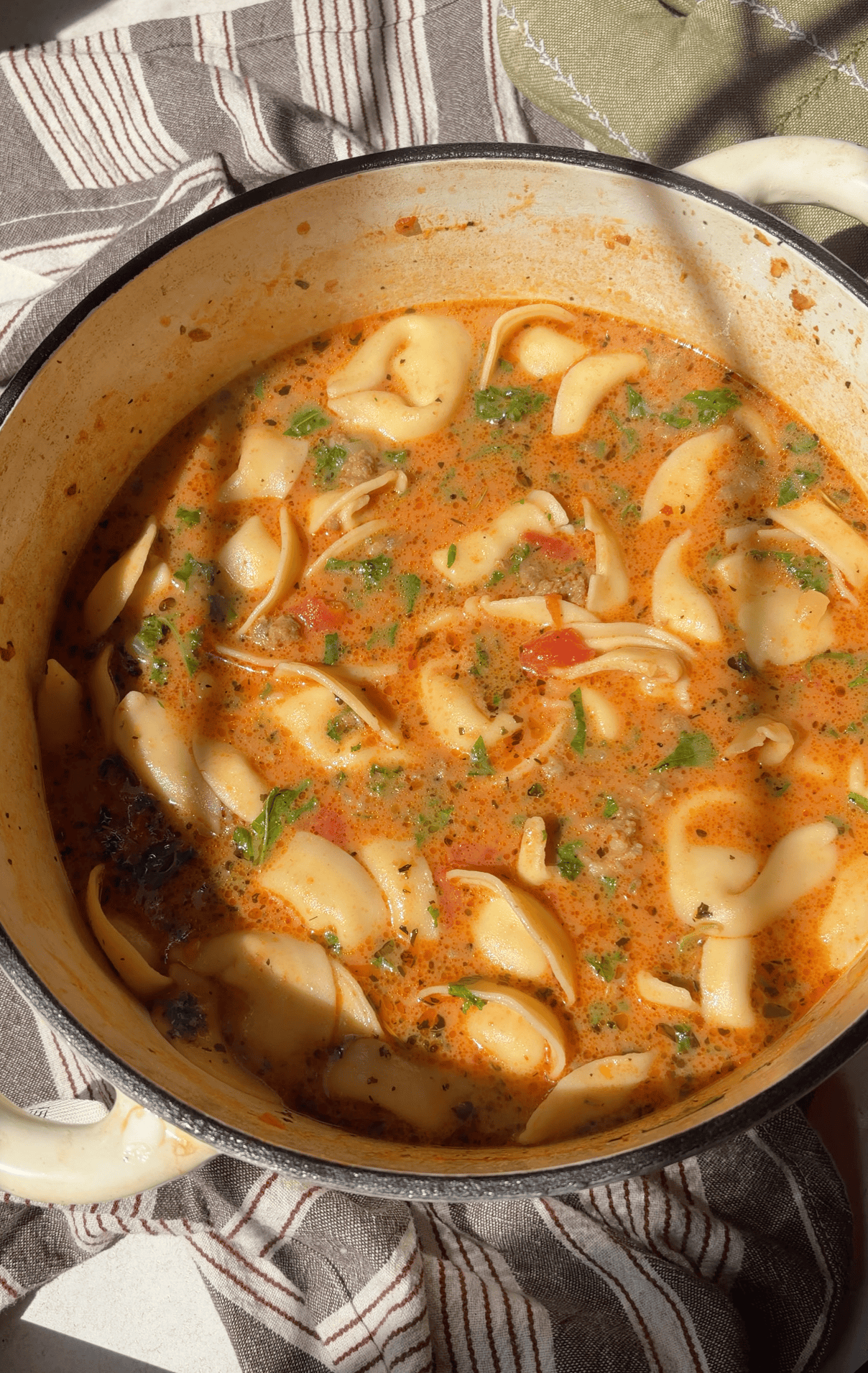 A white pot filled with creamy sausage tortellini soup with herbs, sits on a striped cloth with a green oven mitt nearby. Sunlight highlights the rich colors of the dish.