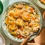 A bowl of creamy slow cooker chicken pot pie filling with carrots, peas, corn, and green beans, topped with golden, flaky biscuit squares. A fork rests beside the food in the floral-patterned bowl.