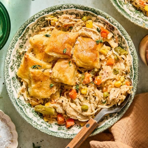 A bowl of creamy slow cooker chicken pot pie filling with carrots, peas, corn, and green beans, topped with golden, flaky biscuit squares. A fork rests beside the food in the floral-patterned bowl.