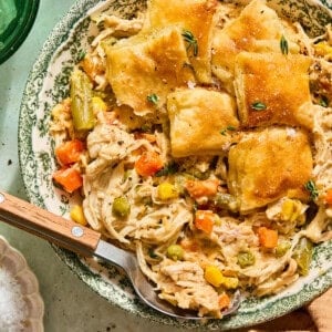 A bowl of chicken pot pie with golden biscuit crust, shredded chicken, carrots, peas, corn, and green beans. A spoon rests inside the bowl, and fresh herbs are sprinkled on top.