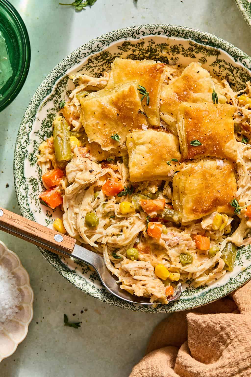 A bowl of crock pot chicken pot pie with golden biscuit crust, shredded chicken, carrots, peas, corn, and green beans. A spoon rests inside the bowl, and fresh herbs are sprinkled on top.