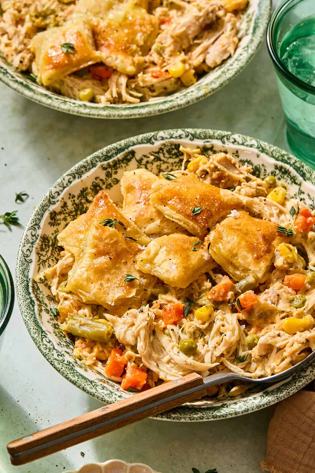 A bowl of crock pot chicken pot pie with golden biscuit pieces with a fork. Another similar bowl and a green glass of water are partially visible nearby.