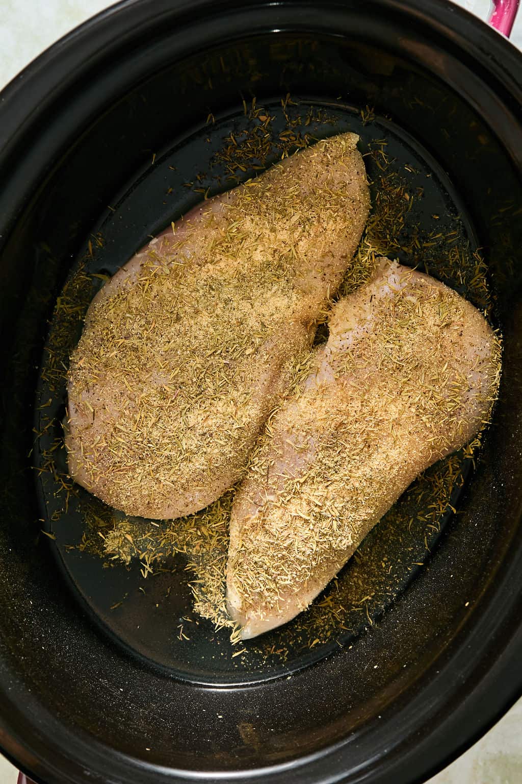 Raw, seasoned chicken breasts with dried herbs in a black slow cooker, ready to be cooked.