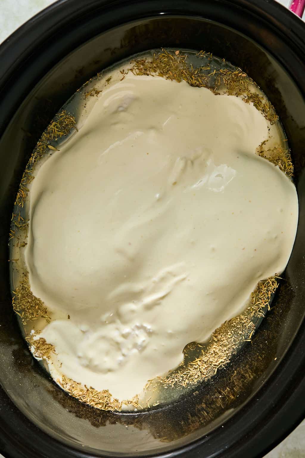 A close-up of a slow cooker with cottage cheese and chicken bouillon paste and dried herbs spread on the bottom, ready for cooking.