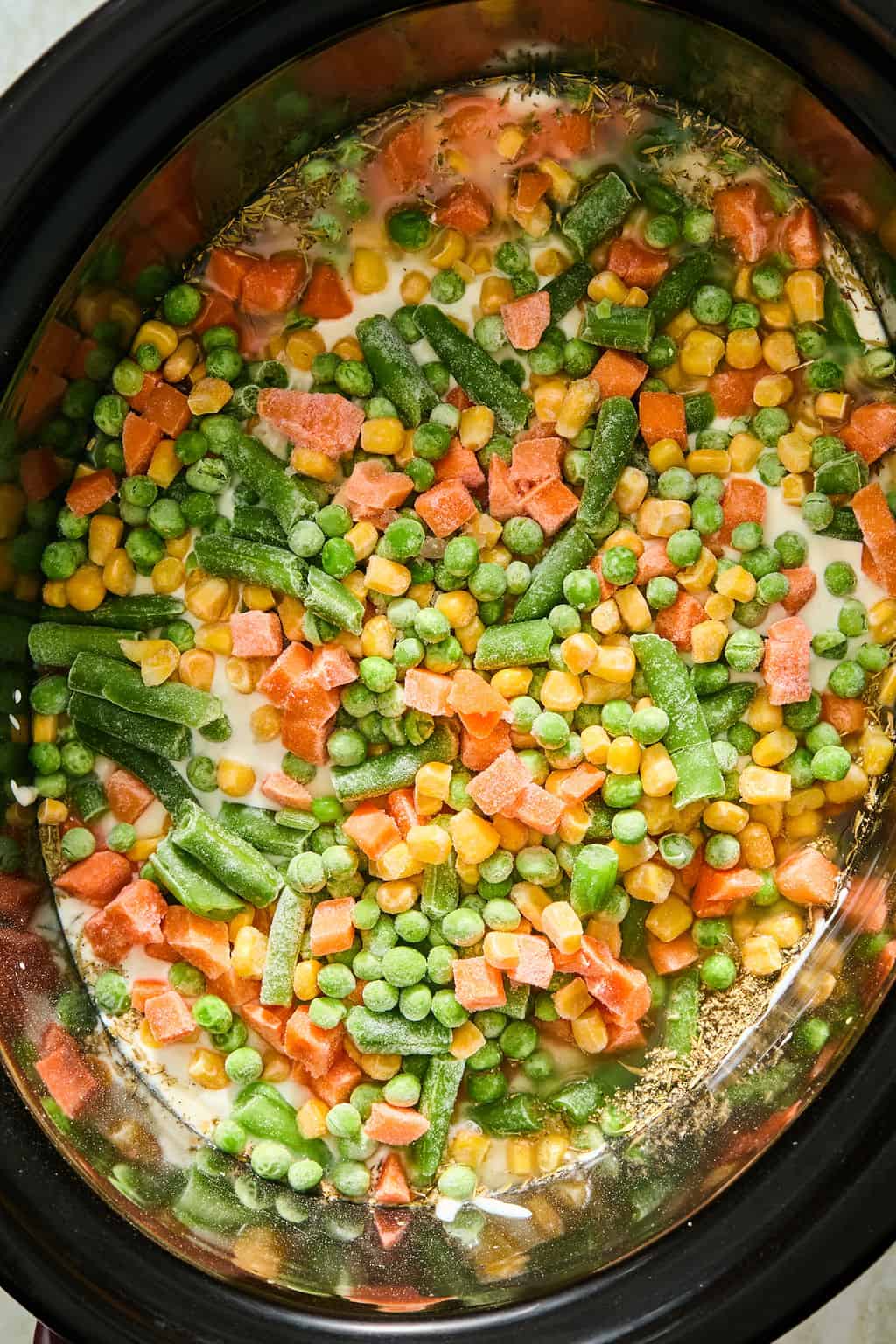 A slow cooker filled with a mixture of frozen vegetables, including peas, carrots, corn, and green beans, in a creamy liquid.