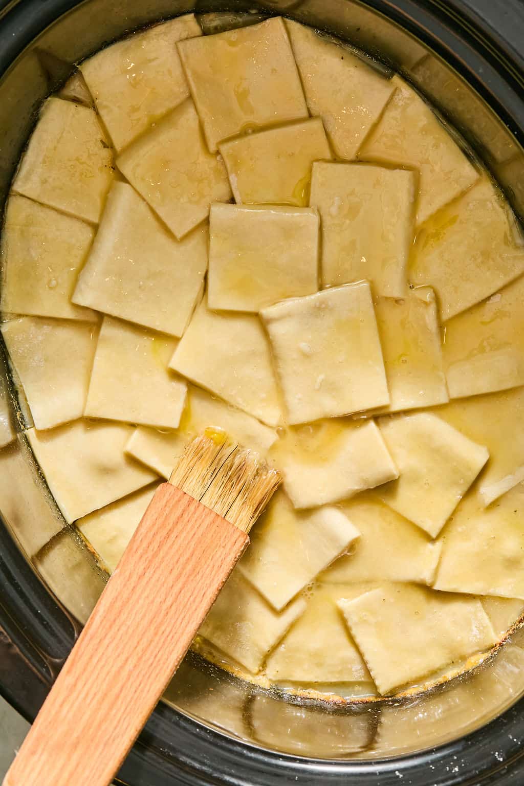 Uncooked square puff pastry pieces are layered in a slow cooker, coated with whisked egg wash using a wooden pastry brush.