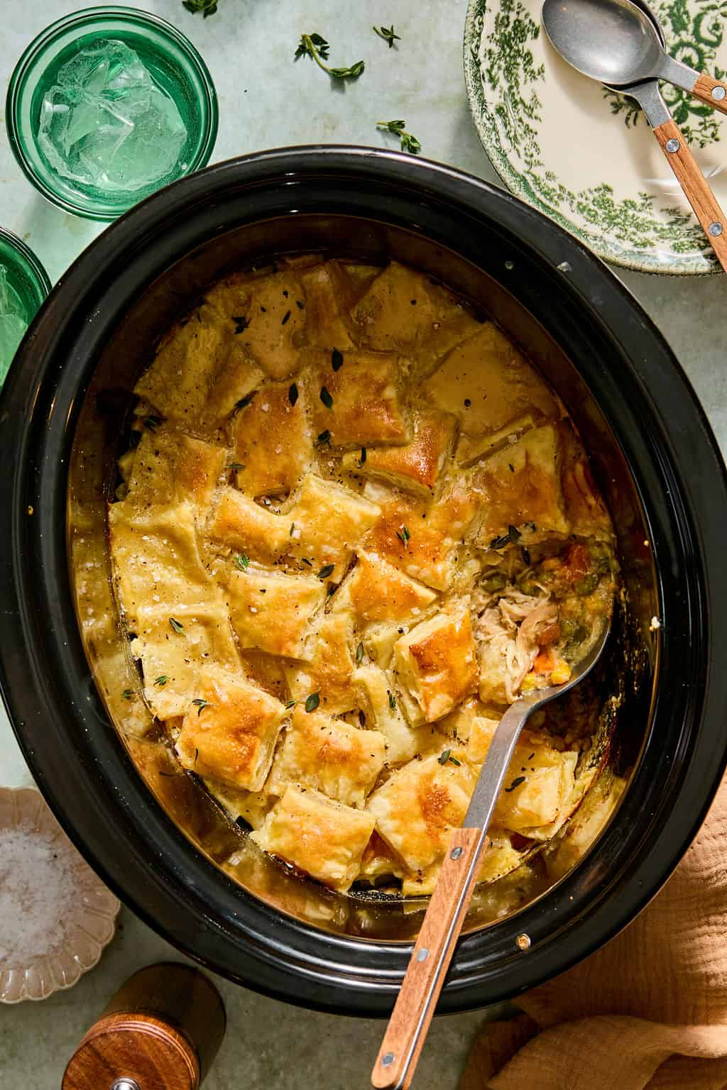 A slow cooker filled with golden-brown, square biscuit-topped chicken pot pie, with a serving spoon scooping some out. Two green glasses, a floral plate, and utensils sit nearby.