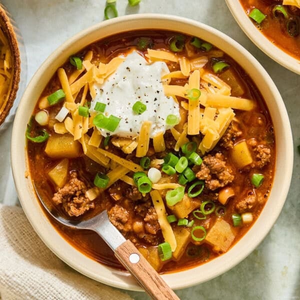 A bowl of hearty smashburger soup, topped with shredded cheddar cheese, sliced green onions, and a dollop of sour cream, with ground meat and diced potatoesโspoon resting inside.