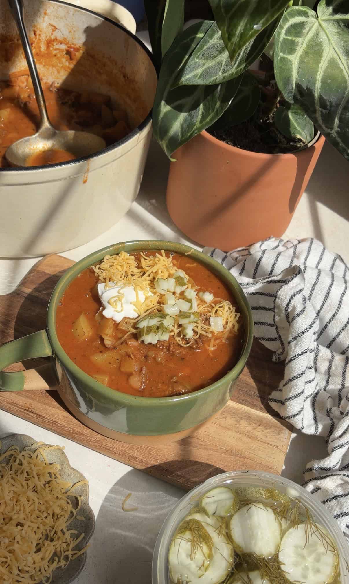 A bowl of smashburger soup topped with shredded cheese, onions, and sour cream sits on a wooden board next to a pot of soup, a striped cloth, and a plant in sunlight.