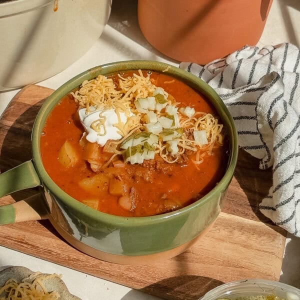 A bowl of hearty smashburger soup topped with shredded cheese, sour cream, diced onions, and herbs sits on a wooden board, beside a striped cloth napkin.