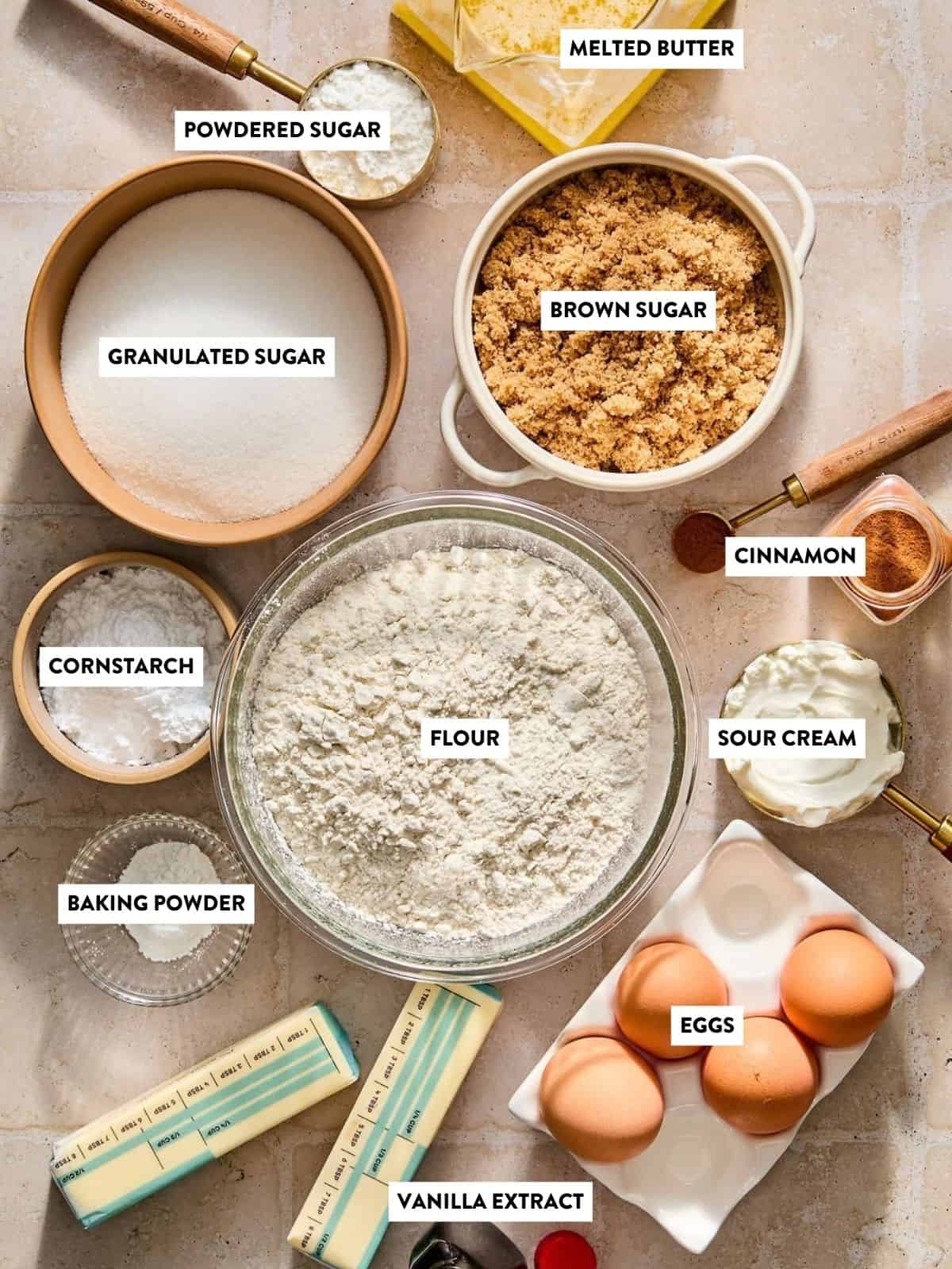 Top view of baking ingredients for sour cream coffee cake including flour, eggs, granulated sugar, brown sugar, powdered sugar, sour cream, butter, cornstarch, baking powder, cinnamon, and vanilla extract, each labeled in bowls on a countertop.