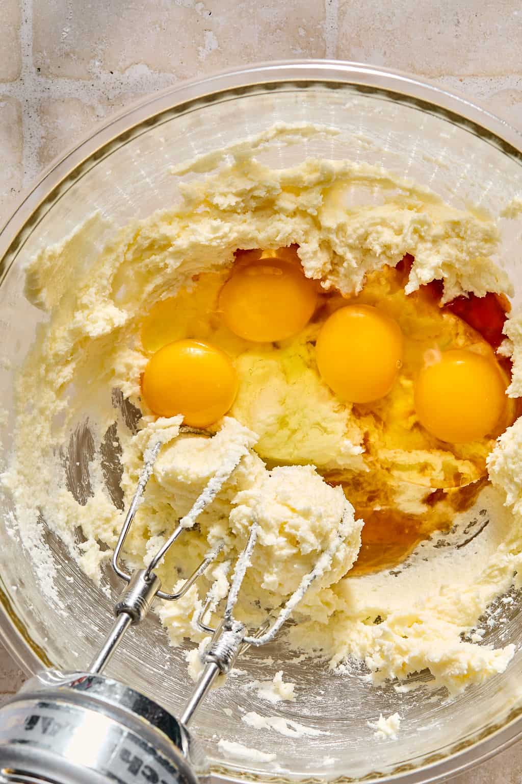 A glass bowl with creamed butter and sugar, four raw eggs on top, and a hand mixer with beaters partially covered in batter, set on a light-colored countertop.