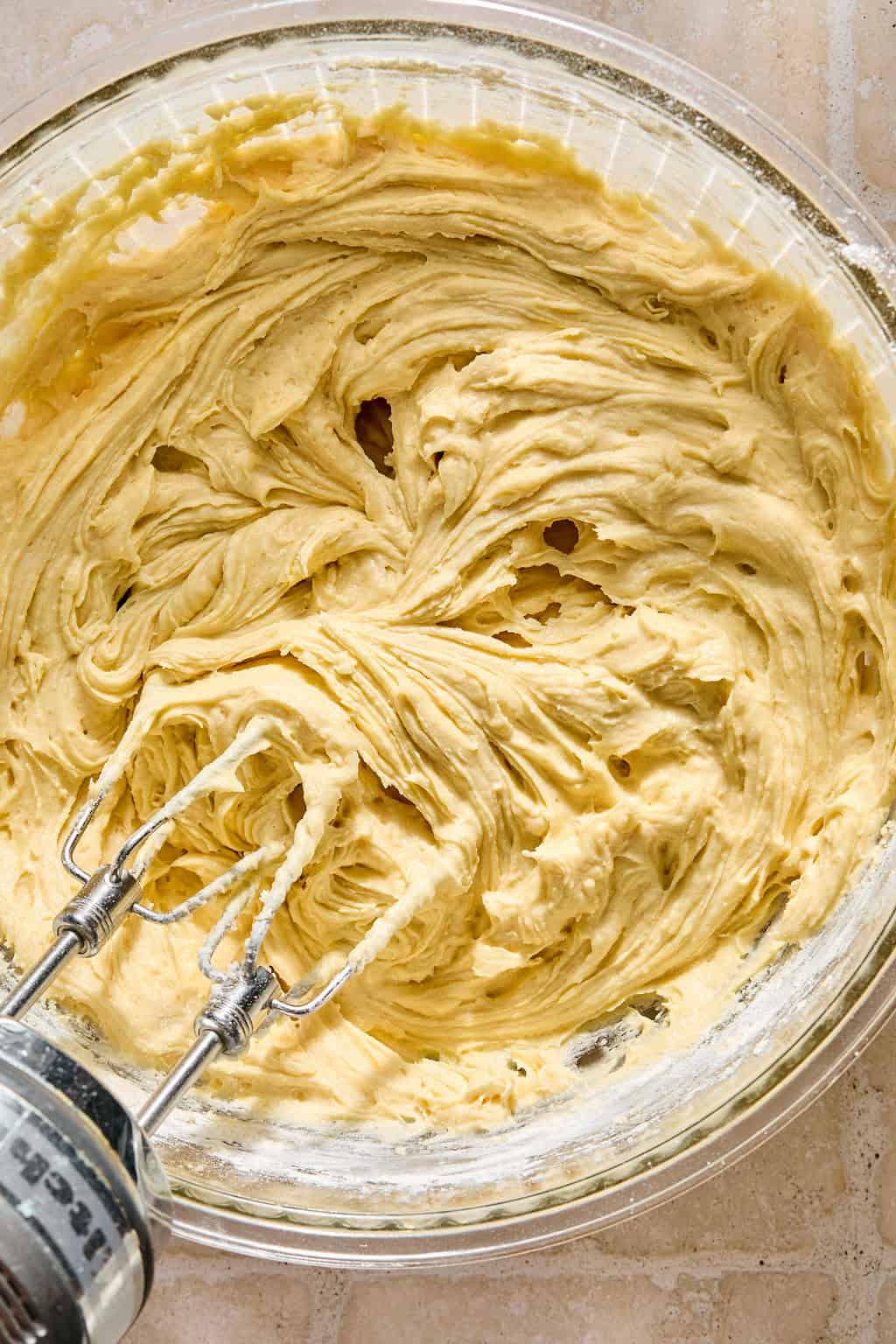A glass bowl filled with creamy yellow cake batter being mixed by an electric hand mixer with beaters, resting on a light-colored countertop.