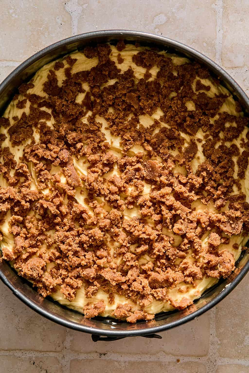 A round baking pan filled with unbaked coffee cake batter, topped with a layer of crumbly brown streusel, sits on a light-colored tiled surface.