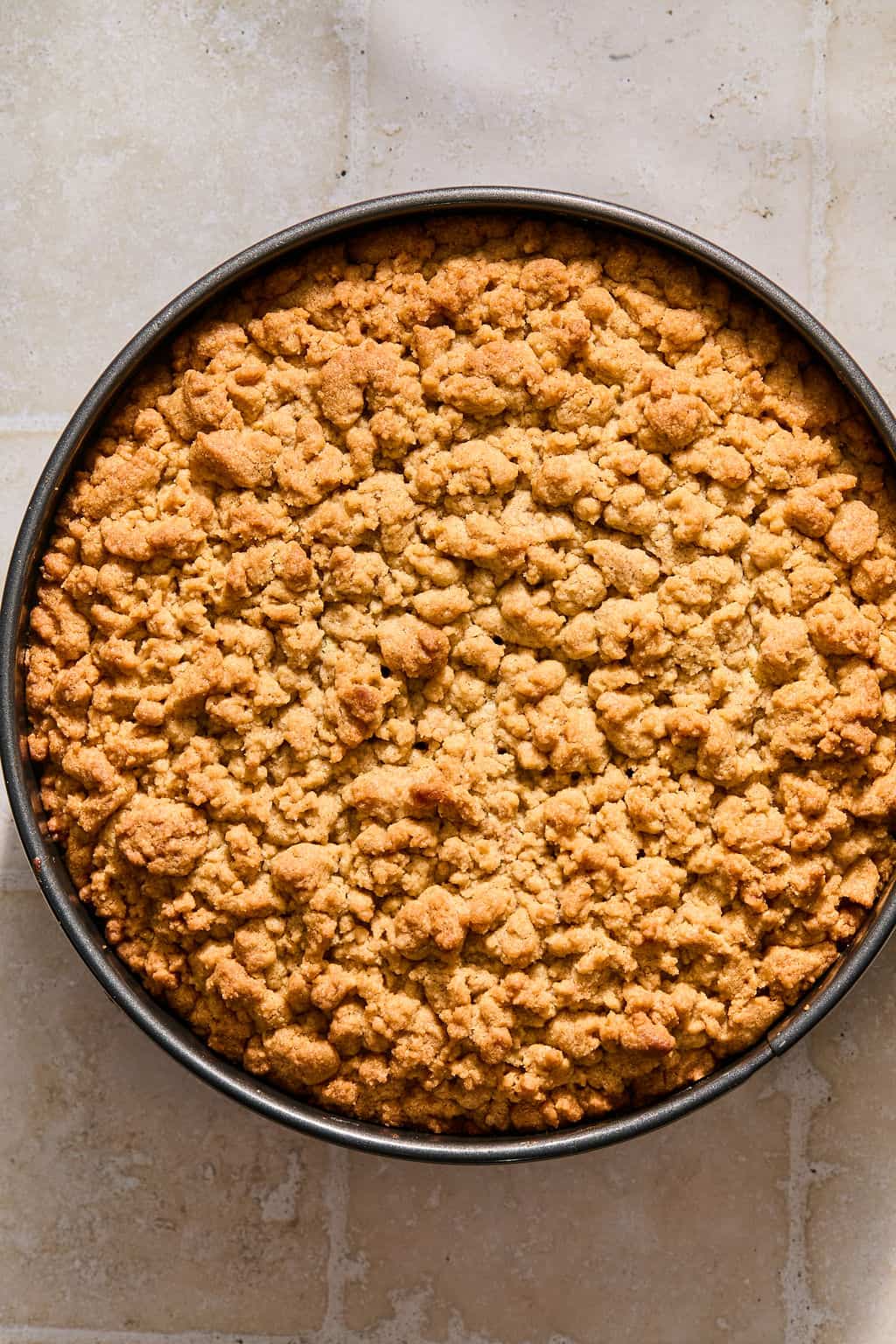 A round crumble cake with a golden, crumbly topping sits in a metal pan on a light tiled surface.