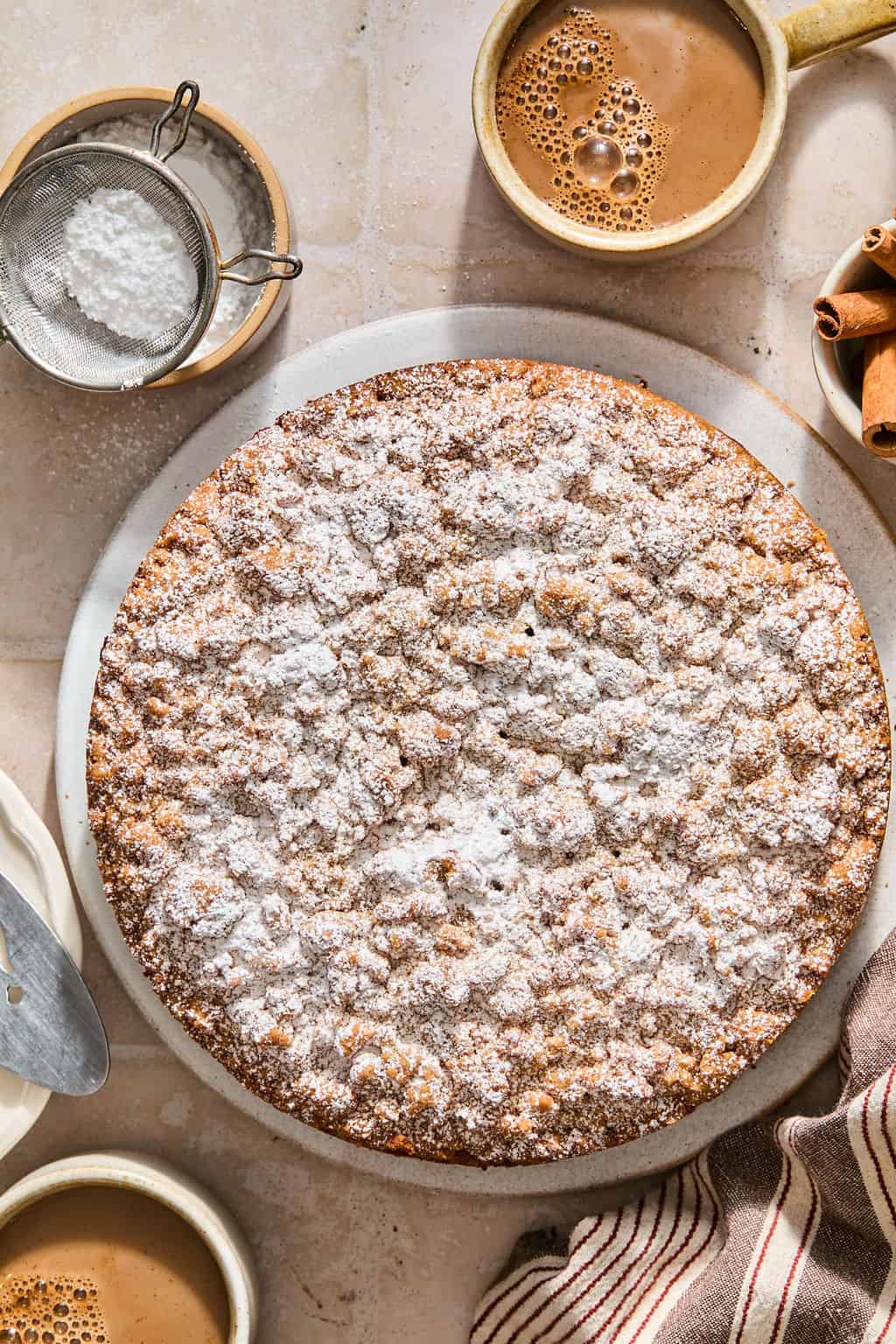 A round sour cream coffee cake dusted with powdered sugar sits on a marble board, surrounded by cups of coffee, a strainer with sugar, cinnamon sticks, and a striped cloth.