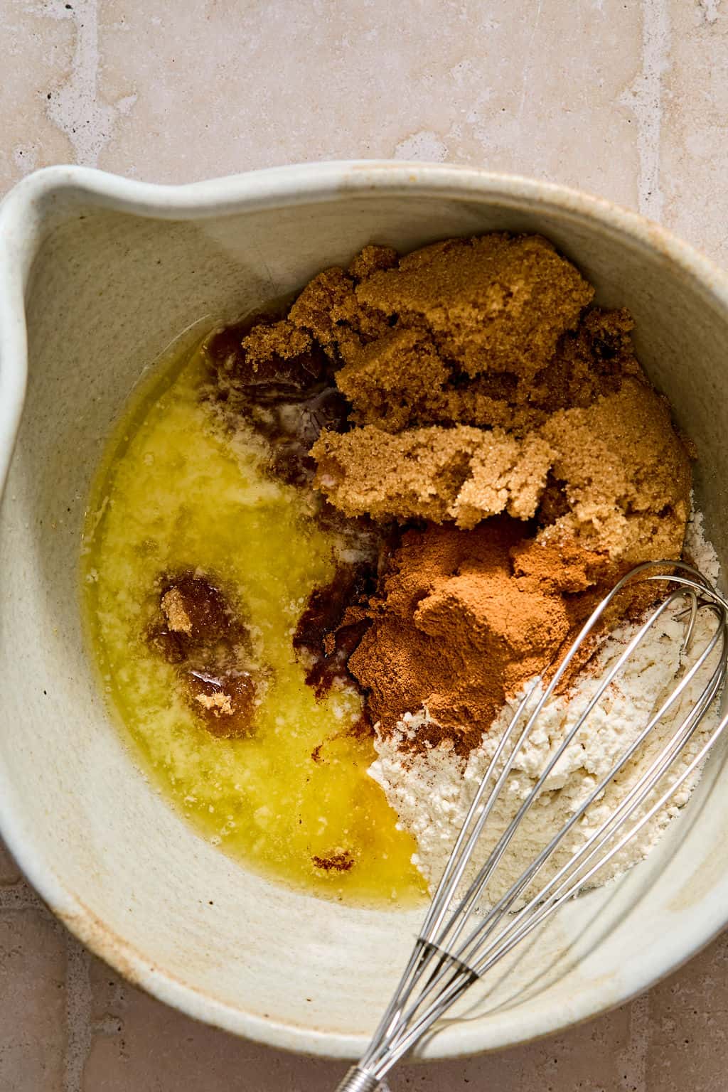 A mixing bowl with melted butter, brown sugar, flour, and cinnamon, next to a metal whisk on a light-colored surface.