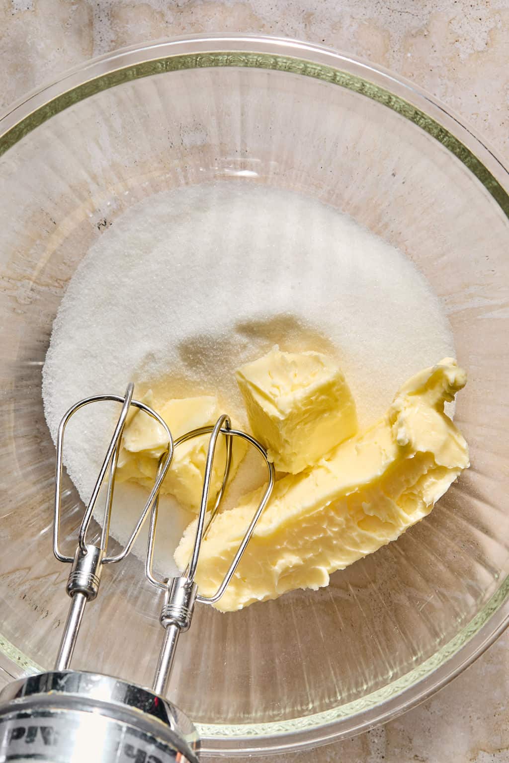 A glass bowl with sugar and butter, ready to be mixed with a hand mixer on a light countertop.