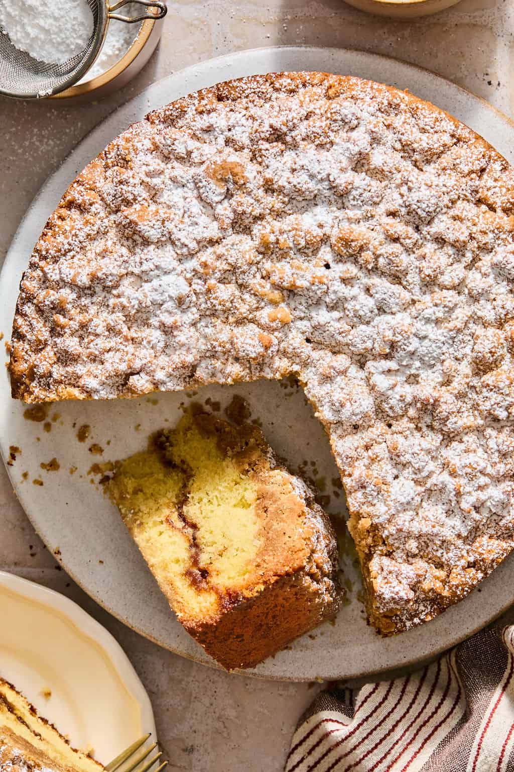 Round sour cream coffee cake dusted with powdered sugar sits on a plate, with one large wedge cut out and placed nearby, revealing a swirled cinnamon layer inside. A striped cloth and plates are partially visible beside the cake.