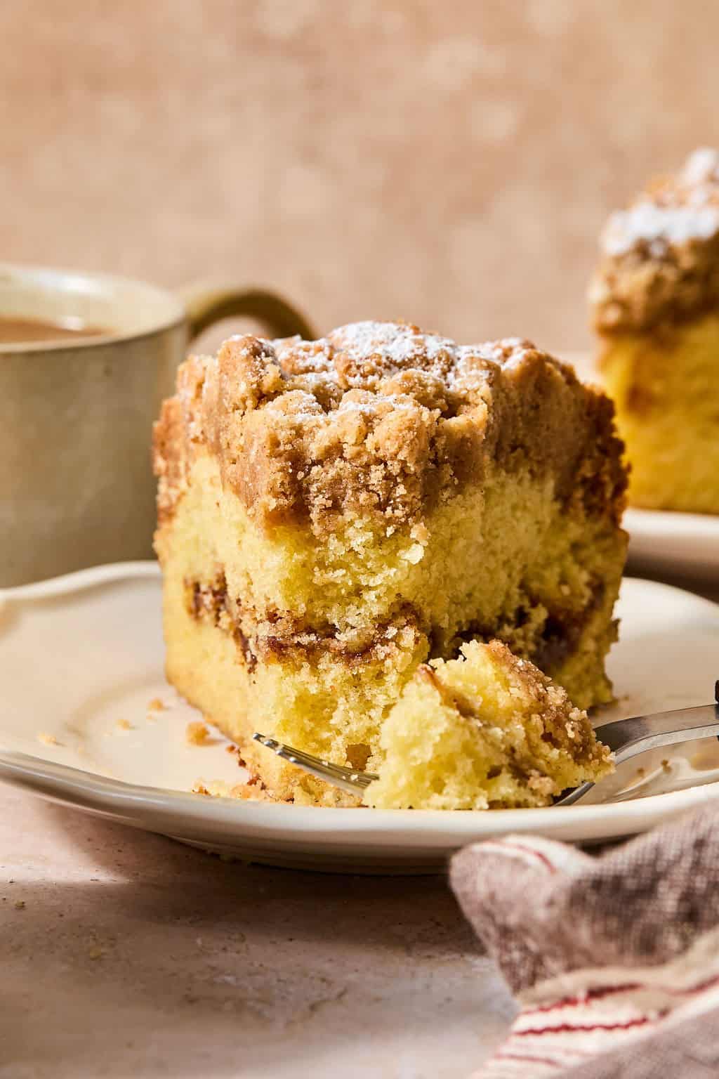 A fork cutting into a thick slice of crumb-topped sour cream coffee cake on a white plate, with a mug and another cake slice in the background.