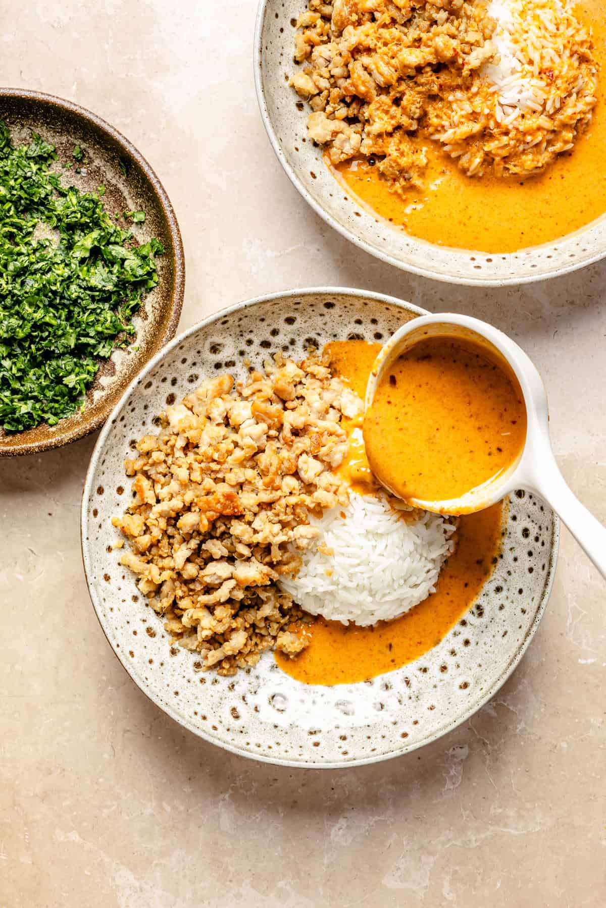 A bowl of white rice topped with ground chicken, being drizzled with a creamy orange curry sauce from a ladle, next to a small bowl of chopped fresh herbs.