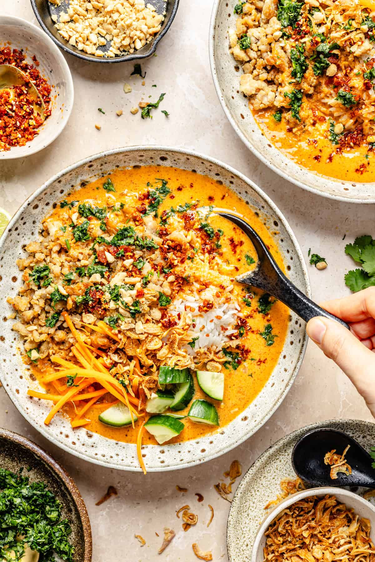 A hand uses a spoon to stir a creamy orange coconut curry broth bowl served with rice, sliced cucumbers, shredded carrots, crushed peanuts, and chili crunch.