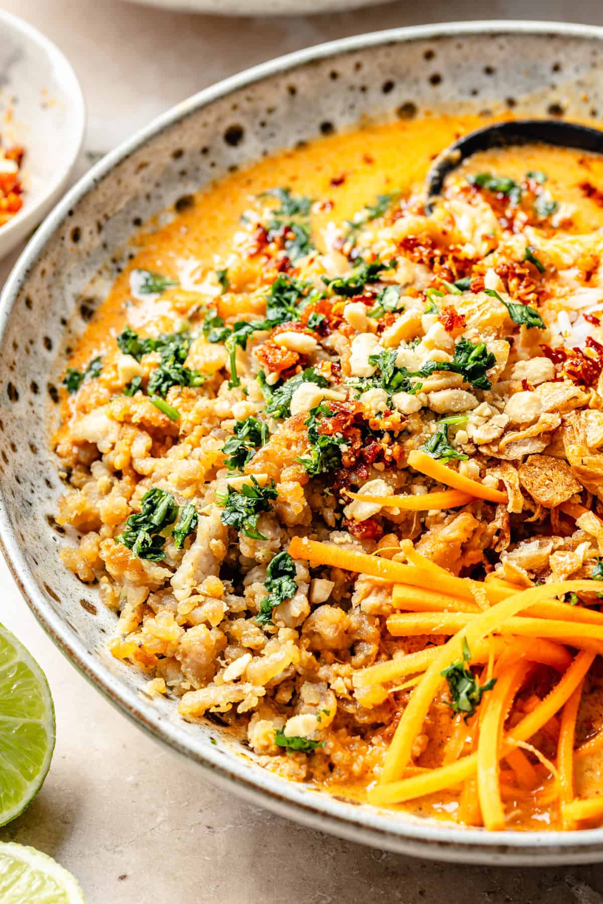 A bowl of Thai-style coconut curry broth bowl topped with crispy shallots, shredded carrots, chopped herbs, and crushed peanuts, with a lime wedge on the side.