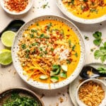 A bowl of coconut curry broth with rice, topped with shredded carrots, cucumber, herbs, and crispy shallots, accompanied by lime wedges and side bowls of chili flakes and fried onions.
